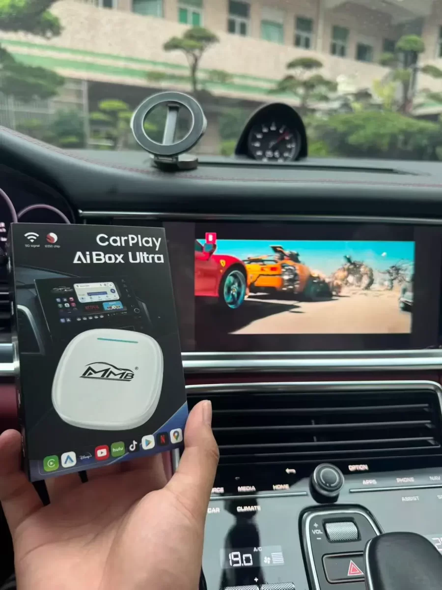 A person holding a CarPlay AiBox Ultra package inside a car, with a display screen showcasing a colorful car racing scene.