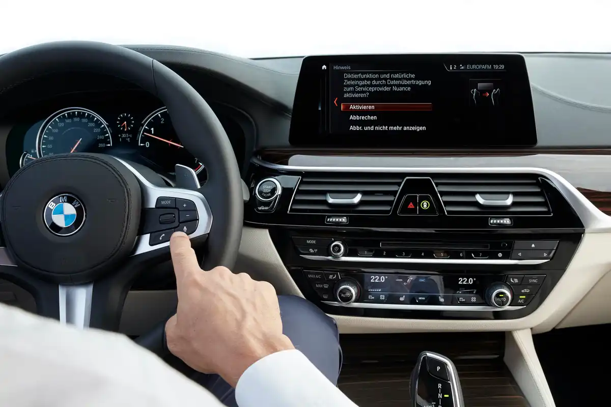 Close-up of a driver's hand pressing a button on a BMW steering wheel, with a display screen showing options in the dashboard.BMW CIC vs NBT vs EVO